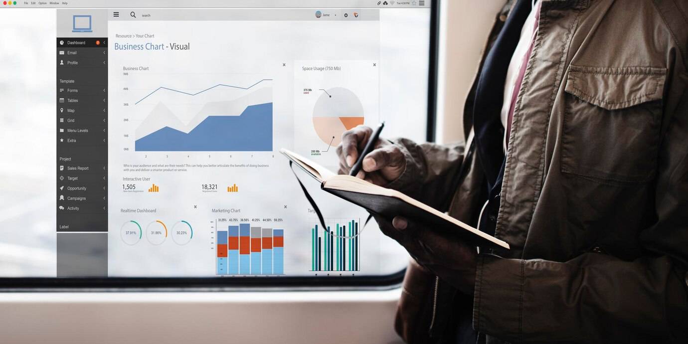 A person writes in a notebook while reviewing a business dashboard displaying charts and analytics on a laptop.