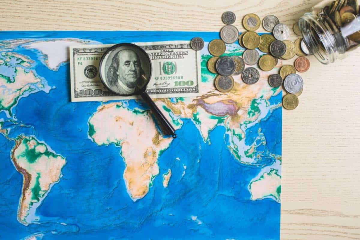 A magnifying glass rests over a $100 bill on a detailed world map, surrounded by scattered coins and a jar of change symbolise the global tax laws.