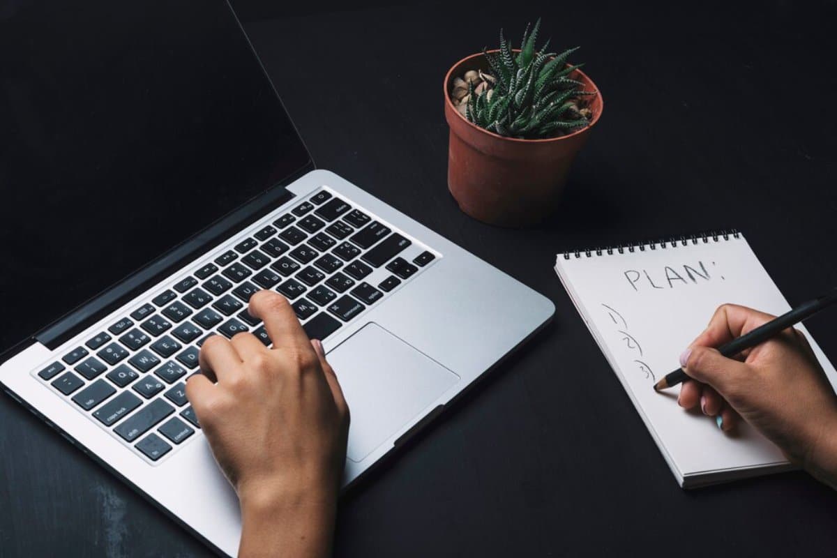 Person using a laptop and writing a plan in a notebook, with a small potted plant beside them.