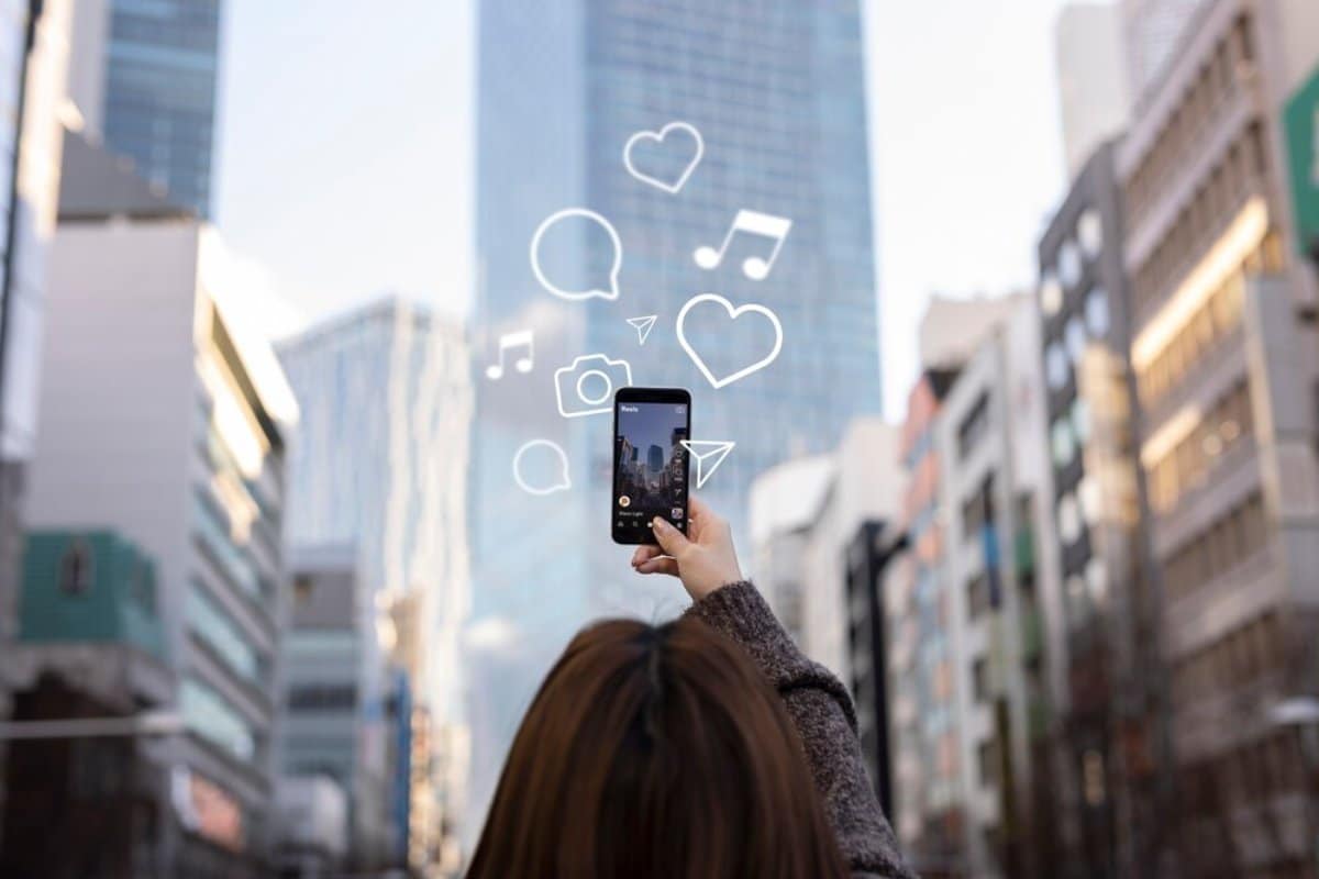 Person holding a smartphone capturing city buildings, with graphical social media icons floating above the phone.