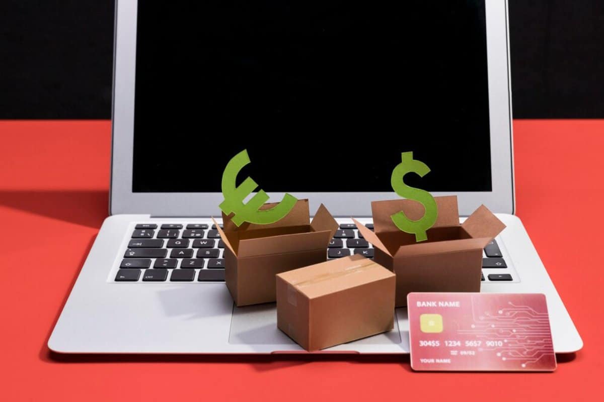 Laptop with currency symbols (euro and dollar) emerging from cardboard boxes, alongside a bank card, on a red and black background.