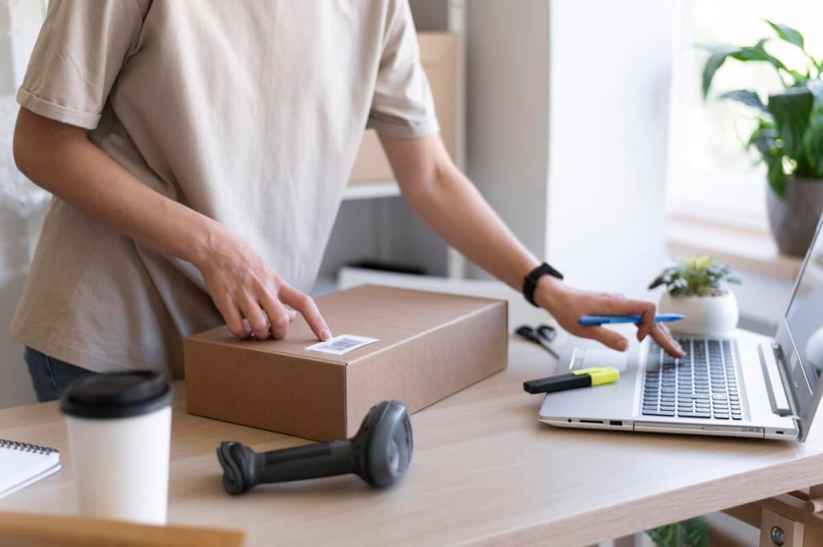 Person shipping parcels, sealing a box at a desk with a laptop, coffee, and shipping tools nearby.