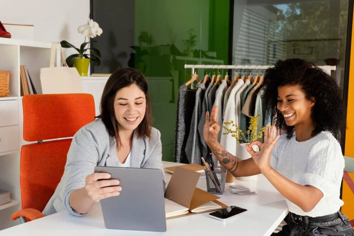 Two women smiling and interacting in a modern office setting, one holding a tablet and the other gesturing a peace sign.
