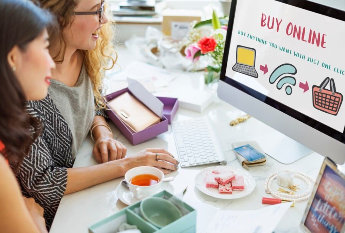 Two women shopping online on a computer, surrounded by jewelry, tea, and snacks, with an on-screen ad promoting online shopping.