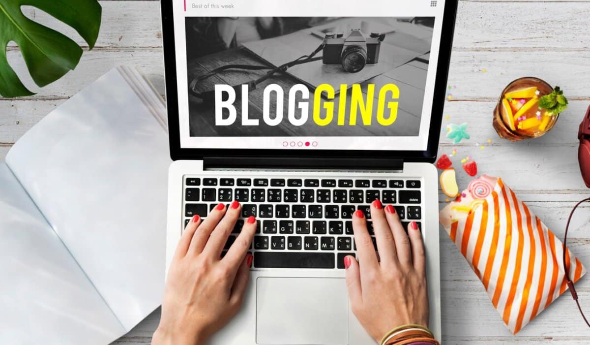 Person using a laptop displaying "BLOGGING" on screen, with snacks and camera nearby on a wooden desk.